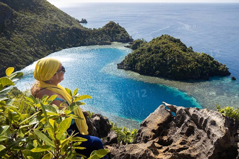 A female tourist enjoys the stunning view of the blue waters and lush islands of Raja Ampat, Indonesia. The vibrant landscape enhances the area's natural beauty