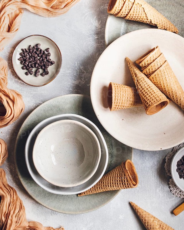 A flat lay of empty waffle cones, ceramic bowls, and a small dish of chocolate chips arranged on a textured surface, offering a rustic and inviting dessert setup