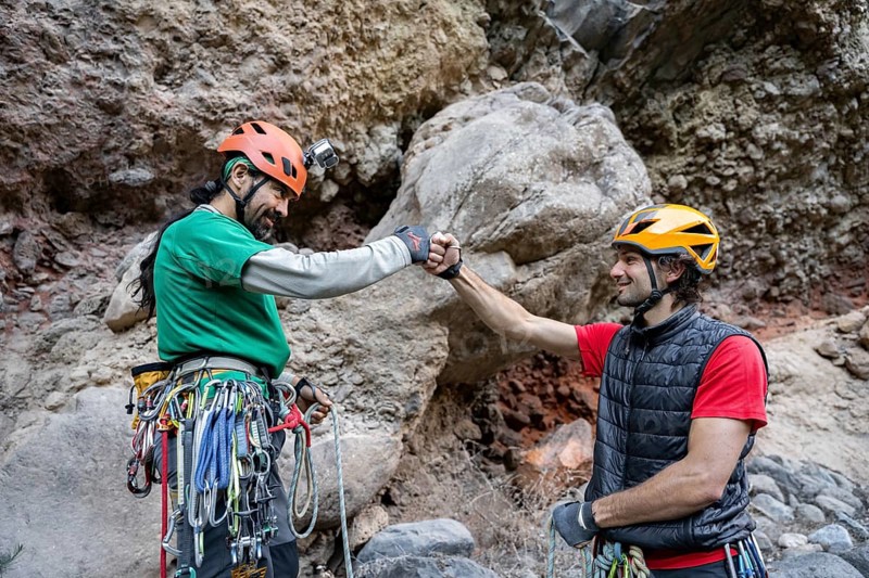 Two climbers, equipped with helmets and climbing gear, share a fist bump in front of a rocky cliff. Their teamwork and camaraderie highlight the spirit of traditional climbing