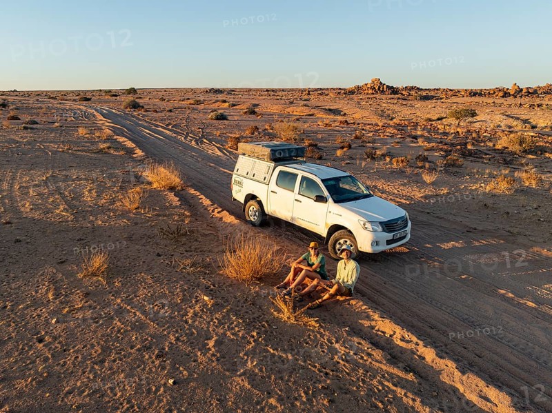 Two travelers sit next to an off-road car on a dirt road in the desert, Erongo, Damaraland, Namibia