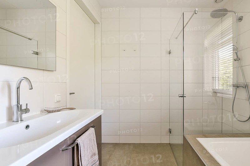 A contemporary bathroom featuring white tiled walls, a glass shower door, and sleek silver fixtures. Natural light floods through a window with blinds, enhancing the minimalist aesthetic