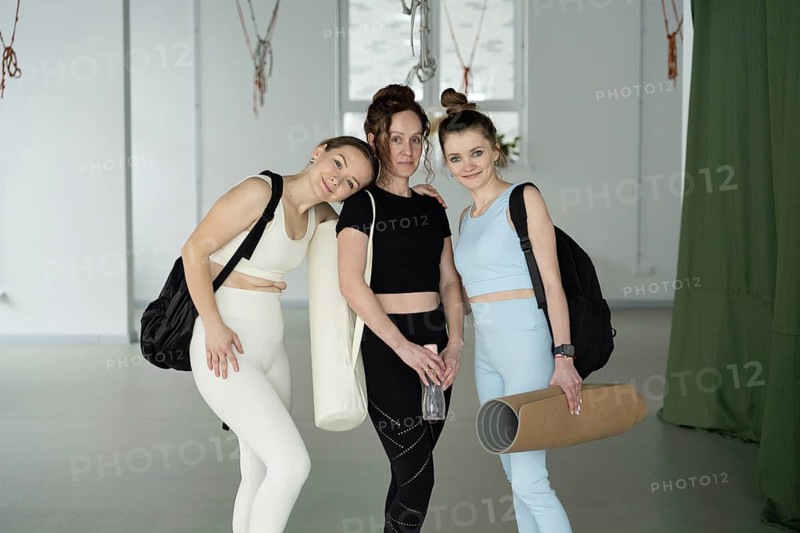 Three women in stylish active wear pose cheerfully in a yoga studio, each carrying mats, embodying fitness and friendship. Ideal image for wellness themes