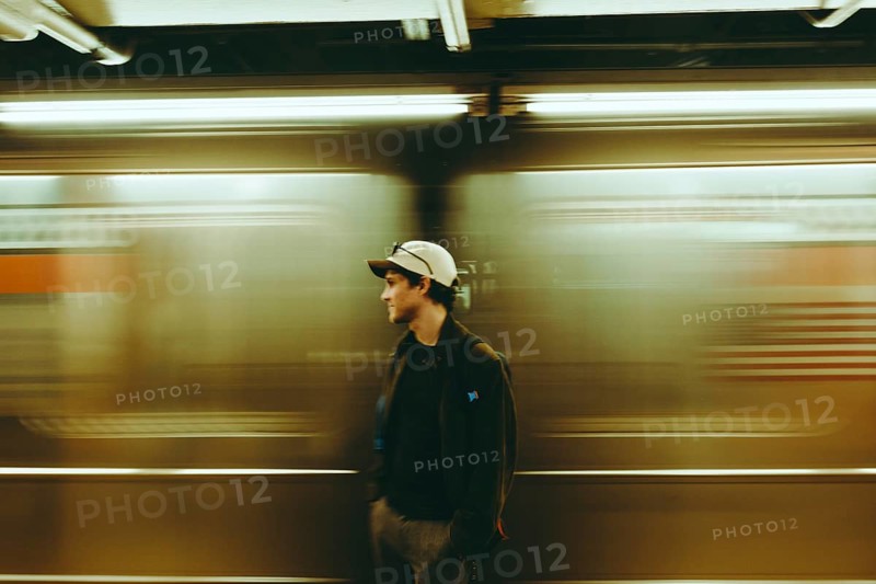 A man wearing a cap stands still on a subway platform while a train speeds by, creating a dynamic blur effect in New York. The scene captures urban travel and modern lifestyle