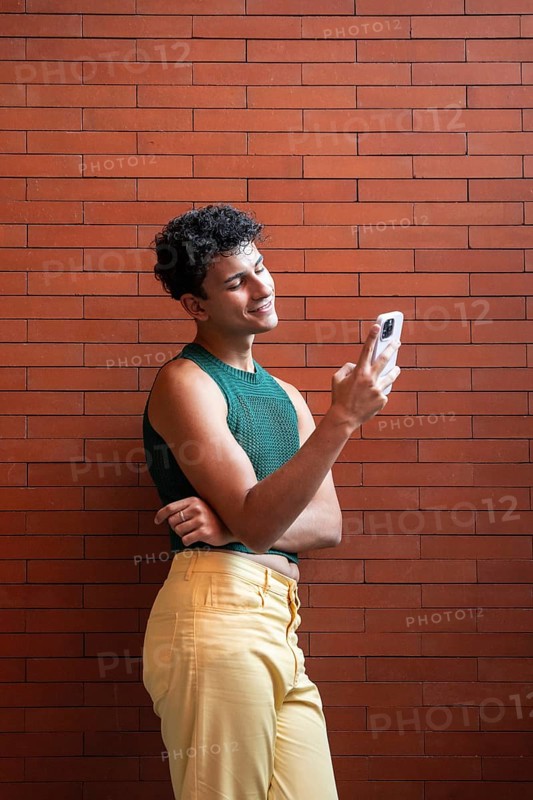 A stylish young person in a green top and yellow pants smiling as they look at their smartphone, standing against a vibrant red brick wall, capturing a joyful moment