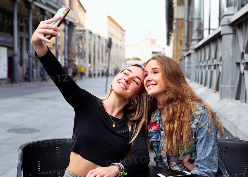 Two teenage girls happily taking a selfie together. They are seated outdoors in a city setting, showcasing their friendship and the joy of capturing moments with a cell phone