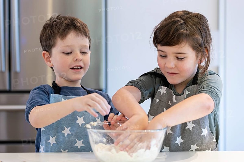 Two children wearing star patterned aprons mix batter in a glass bowl. They smile and engage, showcasing teamwork in a modern kitchen setting, creating joyful family moments