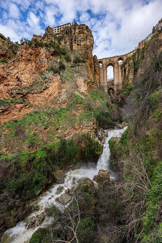 Wide angle view from the bottom of the gorge featuring the massive arches of Puente Nuevo bridge and a small waterfall in Ronda, Spain