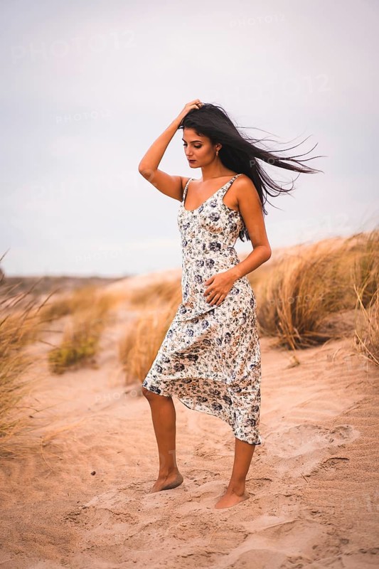Portrait of a Caucasian brunette stroking her hair in a white floral dress, in the sand on the beach, vertical photo