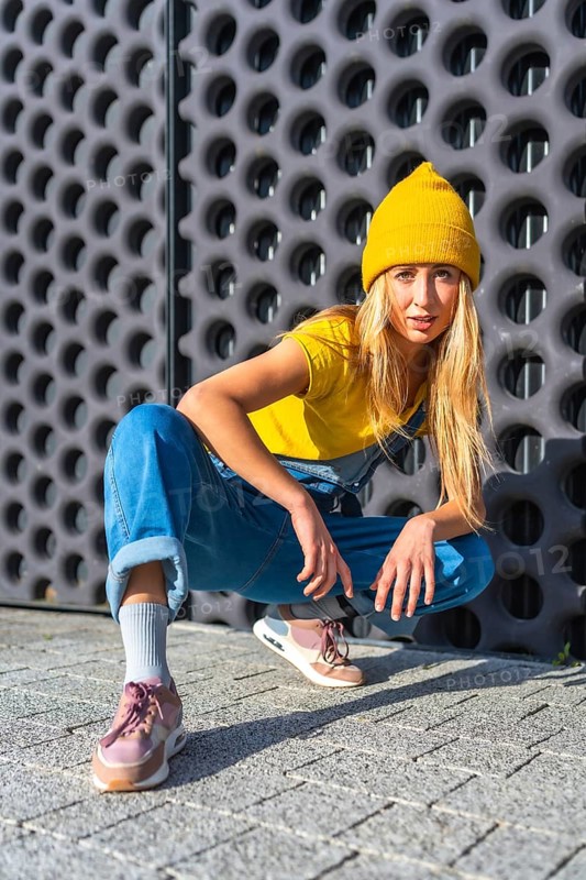 Young woman wearing a yellow beanie, yellow t shirt, and denim overalls, showcasing her urban dance moves against a modern architectural backdrop