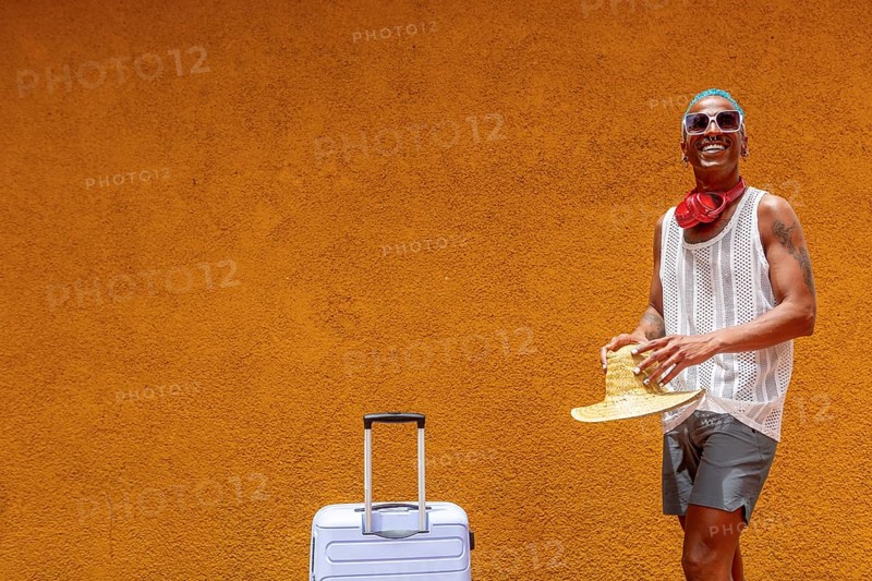 A cheerful LGBTQIA+ man stands with a suitcase against a warm, sunny backdrop. Ready for a holiday, they hold a hat and wear stylish sunglasses and headphones, radiating joy