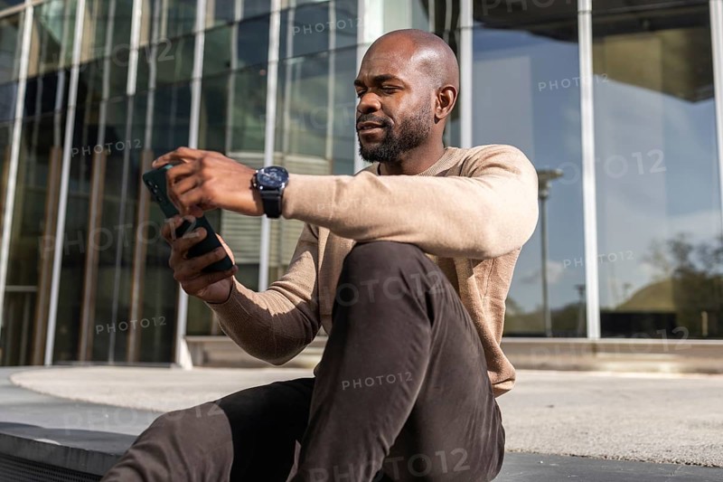 A cuban man sitting outside a modern building, intently looking at his smartphone. He wears a casual outfit with a watch, suggesting a relaxed, tech savvy lifestyle