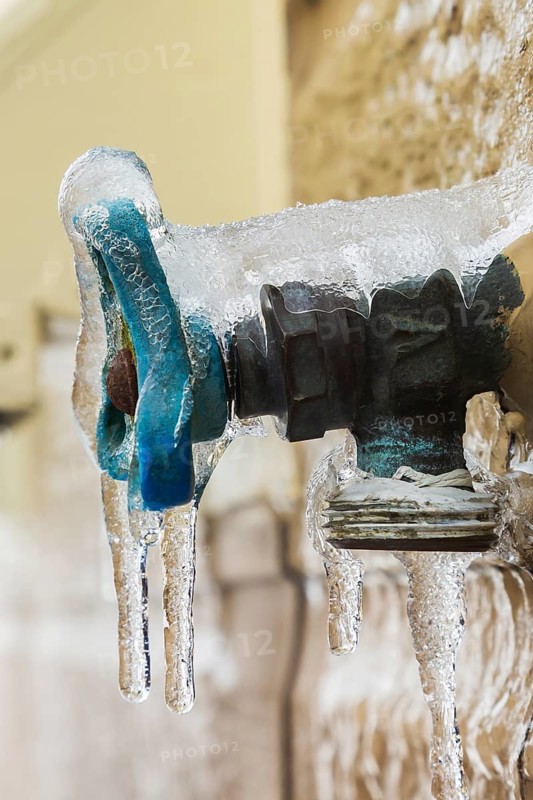 Close-up of outdoor utility brass water faucet with blue handle mounted through Canexel siding on wall of home and covered with ice anf icicles after freezing rain in late winter, Quebec, Canada