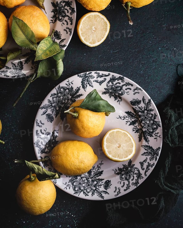 A rustic arrangement of fresh lemons on a plate decorated with lush green leaves. The vibrant yellow color of the lemons contrasts with the intricate design of the plate, creating a visual feast