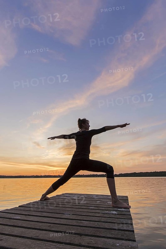 Woman practicing yoga posture Virabhadrasana II, Warrior Pose, lunging standing asana on jetty at lake at sunset