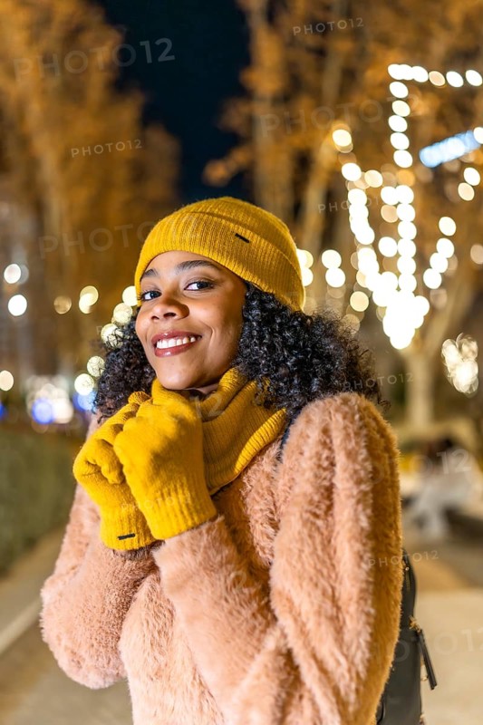 Smiling young woman wearing yellow winter clothes enjoying christmas lights during an evening stroll
