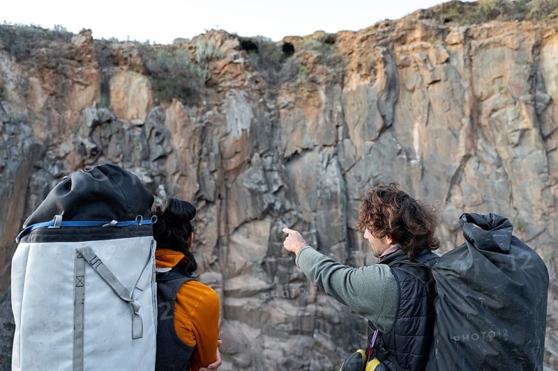 Two climbers equipped with gear discuss their route against a rugged cliff face, embodying the spirit of traditional rock climbing in a natural setting
