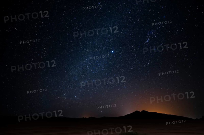 A breathtaking view of a star filled night sky over a Merzouga desert landscape, in Morocco. The silhouetted mountains and a faint glow on the horizon create a serene and mystical atmosphere