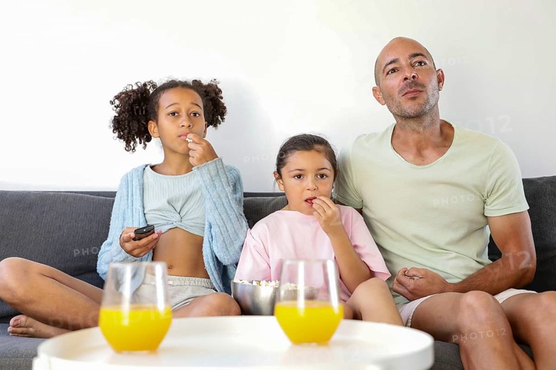 A family sits on a sofa watching a movie together. Two children and an adult enjoy popcorn, with glasses of orange juice on the table, creating a cozy living room atmosphere