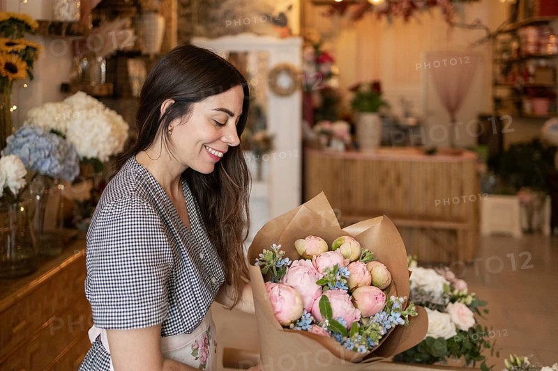 A woman arranges a bouquet of pink and blue flowers in a charming flower shop. She smiles, wearing a checked blouse and floral apron, surrounded by colorful blooms and rustic decor