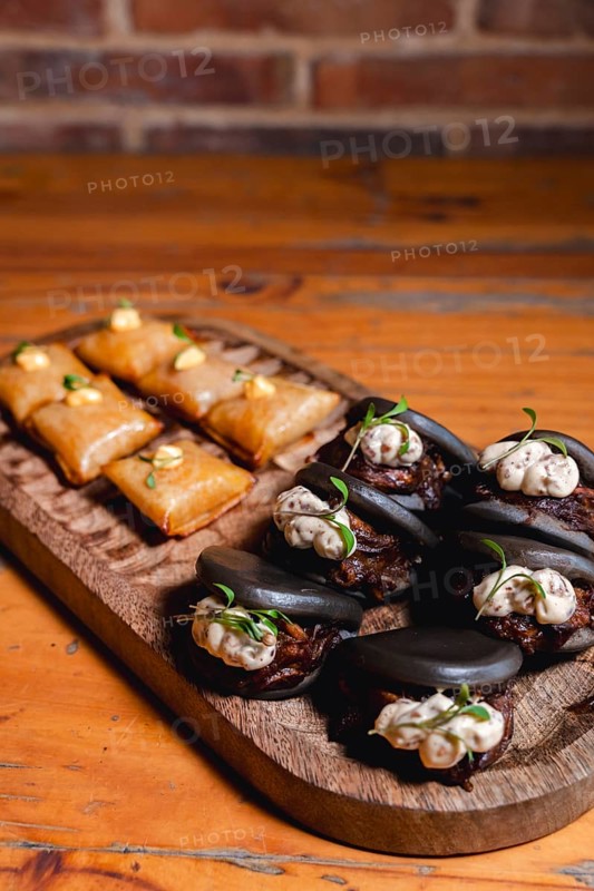 Elegant serving of black octopus bao buns topped with creamy sauce and microgreens, paired with golden-brown appetizers, artfully arranged on a rustic wooden tray over a wooden table