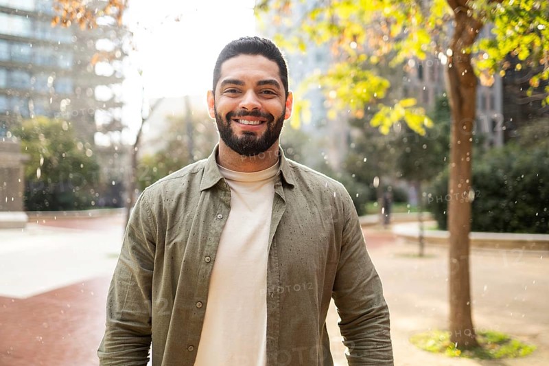 A bearded man smiles warmly under light rain in a park of the city. Sunlight filters through trees, creating a cheerful, peaceful atmosphere. Perfect for themes of joy and serenity