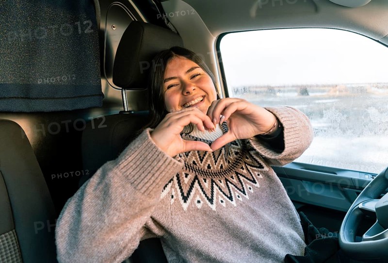 A cheerful Colombian Latina woman in a cozy sweater forms a heart shape with her hands while seated in a vehicle, during a road trip in Iceland. She looks directly at camera with a warm, friendly smile, capturing a moment of happiness