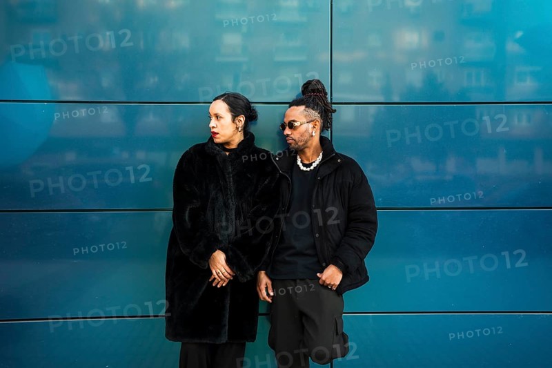 A man and woman from diverse ethnic backgrounds stand against a stylish blue wall. Dressed in modern black attire, they embody multicultural harmony and fashion-forward style