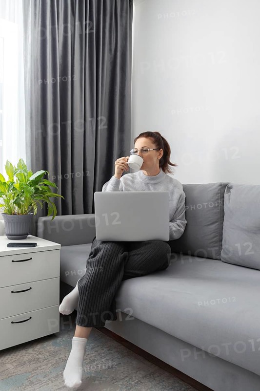 A woman sits comfortably on a sofa, sipping coffee while working on her laptop. She embodies the modern remote work lifestyle, highlighting comfort and productivity