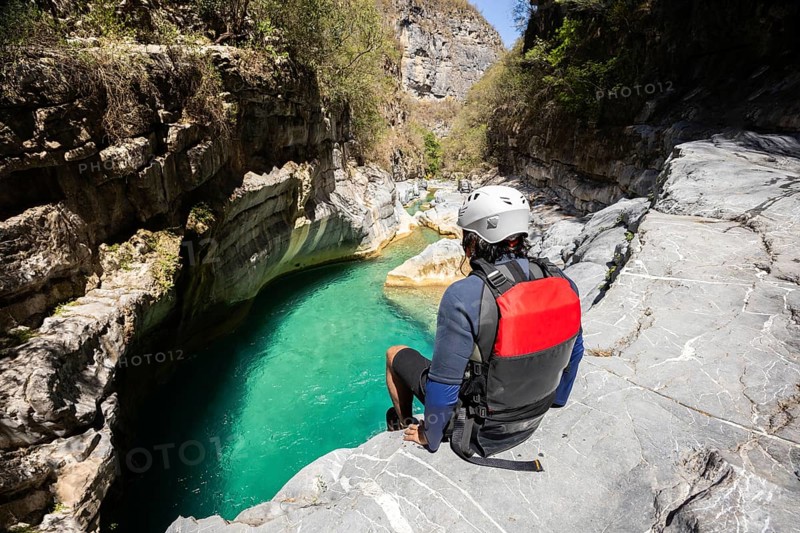 A canyoneer in a helmet and red life vest sits on the edge of a rock cliff, looking down at a beautiful emerald pool within a narrow canyon in Matacanes, Nuevo Leon, Mexico