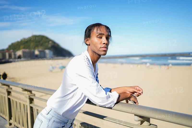 A confident man stands by a beach railing, looking at the camera. The clear blue sky and distant mountain create a serene backdrop, symbolizing pride and individuality