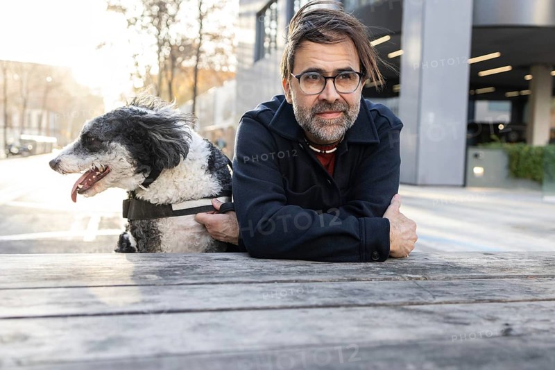 A middle-aged man enjoys a quiet moment with his black and white dog while sitting at a wooden table in an urban park. Both look leisurely and content against the backdrop of a cityscape