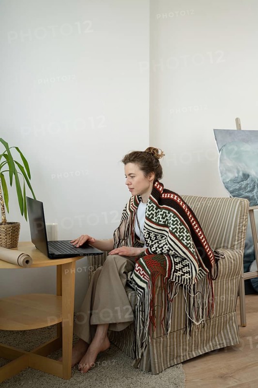 A woman sits comfortably in a striped armchair, wrapped in a patterned blanket. She focuses on her laptop, epitomizing remote work in a stylish, serene setting