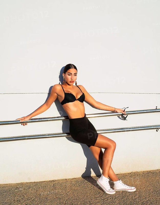 A fashionable woman in black sportswear and white sneakers leans against a railing, holding sunglasses. The outdoor setting and bright sunlight accentuate her bold style