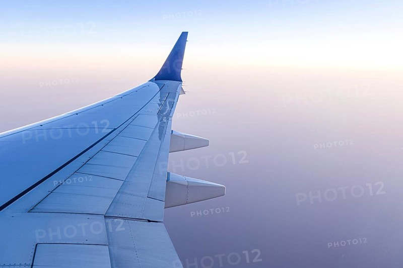 A serene view from an airplane window showing the aircraft wing gliding high above the clouds. The clear sky creates a tranquil and inspiring travel scene