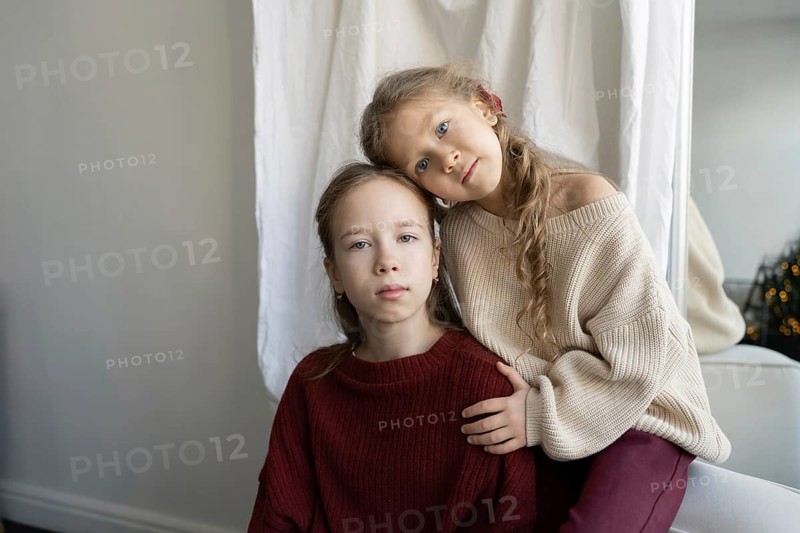 Young sisters pose together, one resting her head on the other's shoulder Both wear cozy, knit sweaters in a tranquil setting, embodying warmth and familial love