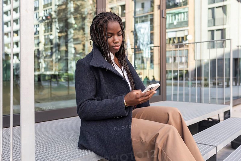 A young African American woman with braided hair, dressed in a business coat, uses a smartphone during a break outdoors