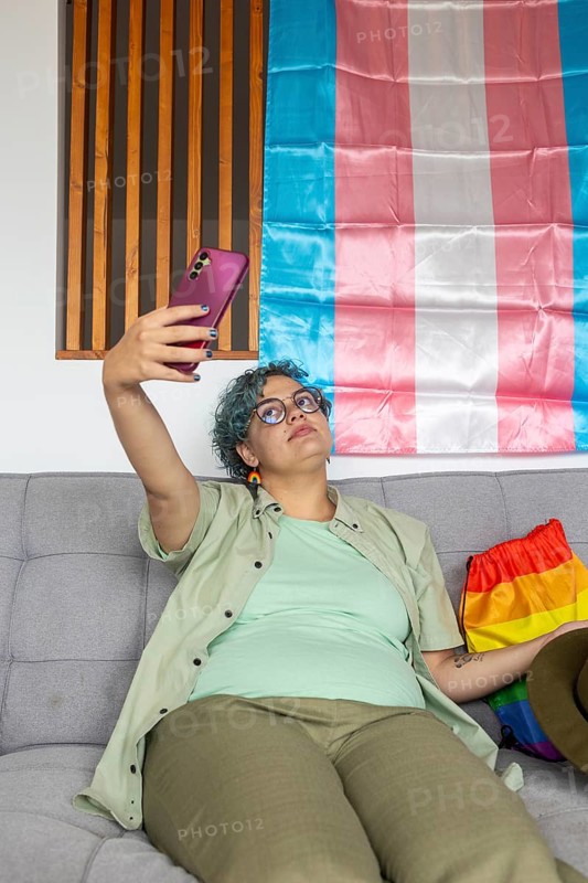 A young transgender person, resting on a couch, takes a selfie with his smartphone in front of a transgender pride flag. He displays a sense of casual comfort and identity expression