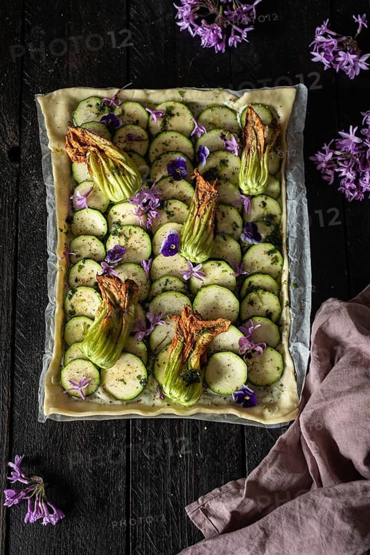 A beautifully arranged zucchini and edible flower tart on a rustic wooden table. The vibrant colors and textures make it an eye-catching culinary masterpiece