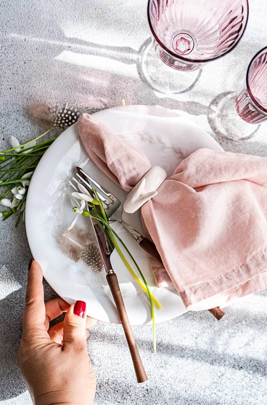 Top view of a cropped unrecognizable woman's hand on an elegant spring table setting featuring snowdrops, a white marble plate, a pink napkin, and vintage utensils. Soft light casts shadows enhancing the delicate arrangement
