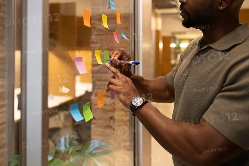 A professional man in a coworking space in Barcelona writes ideas on colorful sticky notes placed on a glass wall, highlighting teamwork and creativity in a collaborative environment