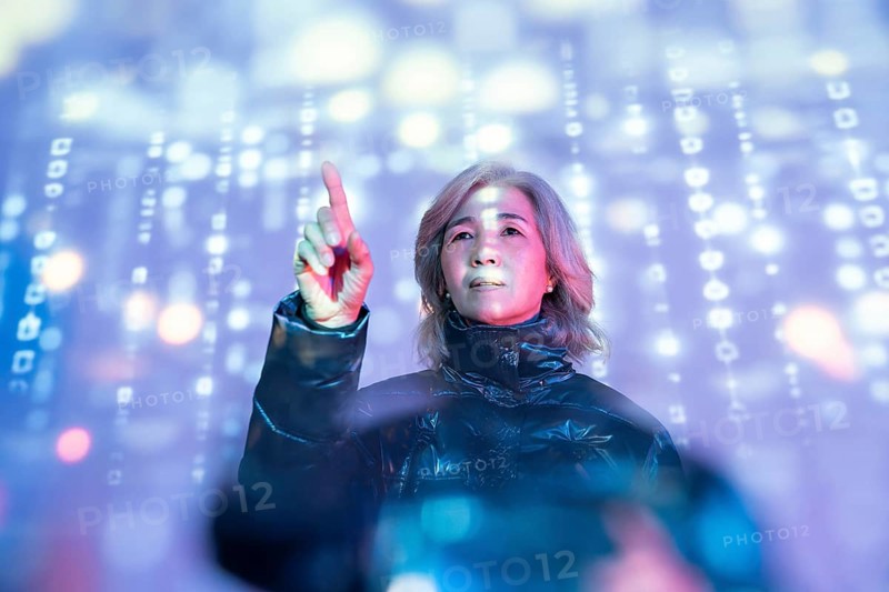 An elderly Asian woman engages with the immersive metaverse, her finger pointing at glowing digital symbols. The vibrant, futuristic setting highlights technology's reach