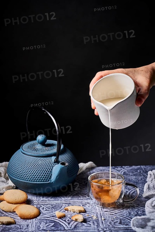 A calming scene with milk being poured into tea from a white jug, surrounded by a traditional teapot and milk cookies on a patterned table. The setup offers a warm, inviting atmosphere