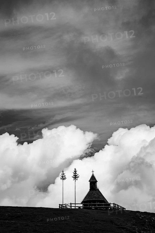 A traditional small wooden chapel with a cross sits on a green grassy ridge under a bright blue sky with large white fluffy clouds in Slovenia