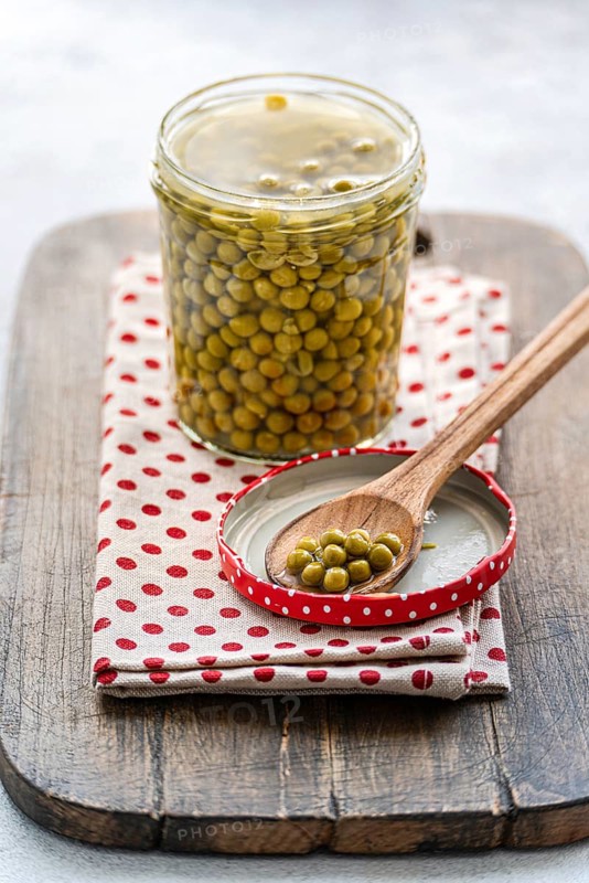 A glass jar of green homemade organic peas displayed on a rustic wooden board, topped with a wooden spoon holding organic peas. The setup includes a red polka dot cloth, creating a homely feel