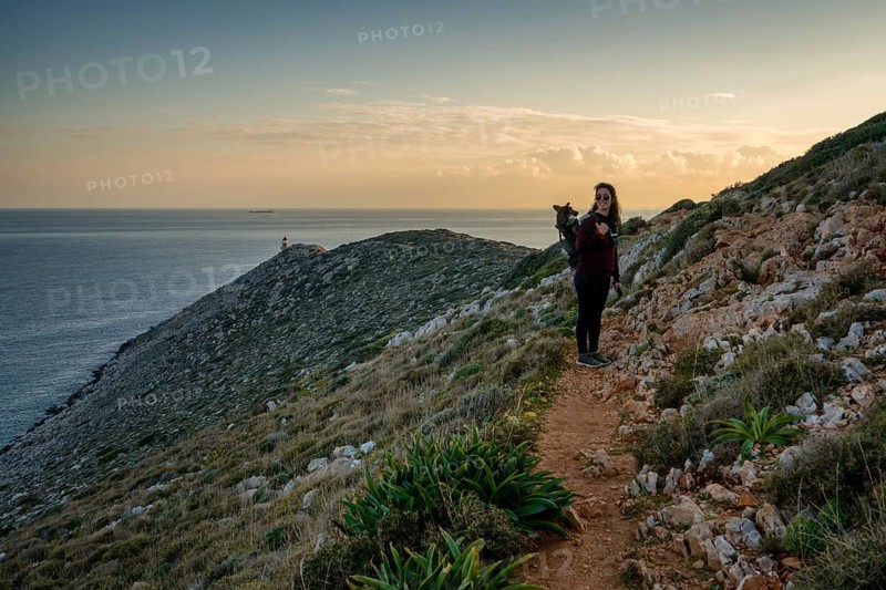 A woman with a dog in a backpack treks along a scenic coastal path in Greece during sunset. The tranquil sea, rocky landscape, and distant Cape Tainaron Lighthouse create a serene setting
