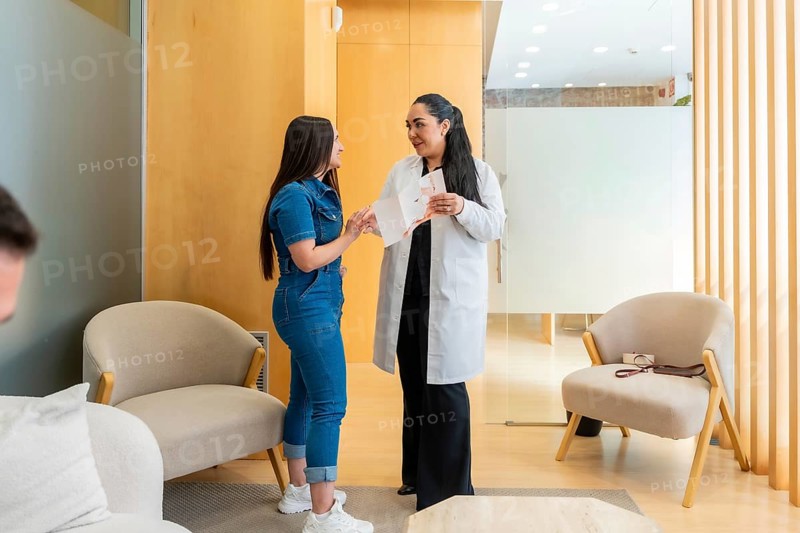 Female doctor in a white lab coat discussing treatment options with a smiling patient wearing a denim jumpsuit in the modern waiting area of an aesthetic or medical center