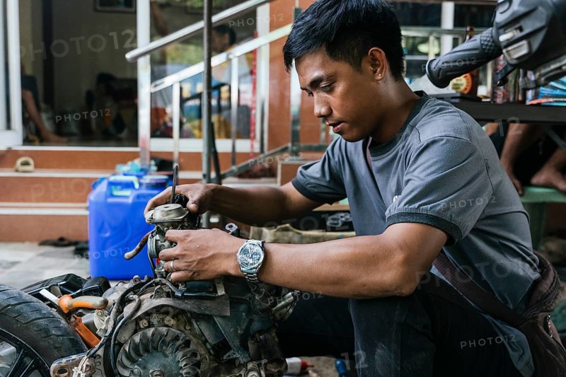 Young mechanic focused on motorcycle engine repair outdoors. Wearing casual attire, he concentrates on a motor part, showcasing skill and dedication in a home based workshop