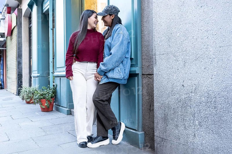 A joyful Latin lesbian couple sharing a loving moment outdoors. They stand closely on a city sidewalk, holding hands, exuding warmth and connection in an urban setting