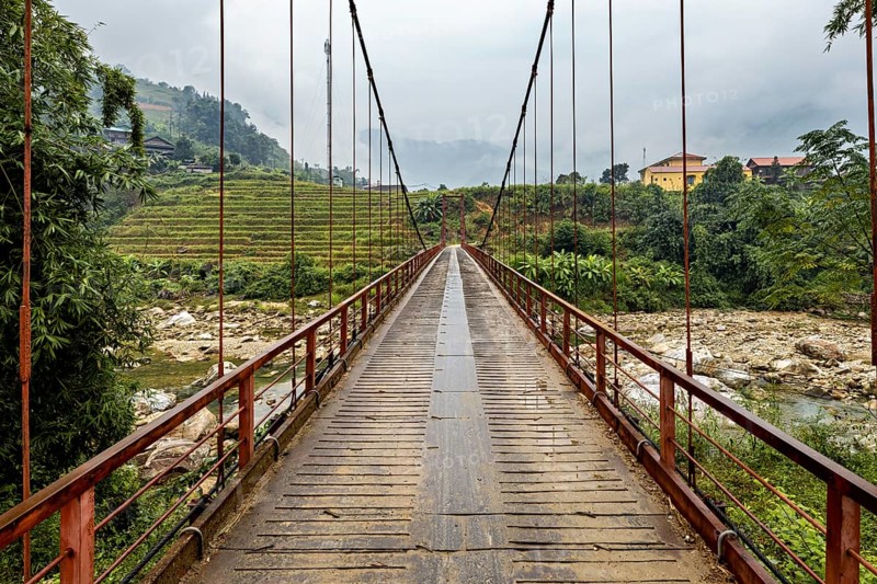 A bridge leads through a tree-lined landscape with rice terraces and ends in the mountains, A bridge over a river in the countryside near Sa Pa in Vietnam