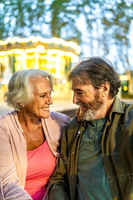 Vertical photo of a modern caucasian senior couple looking at each other sitting on night city fair